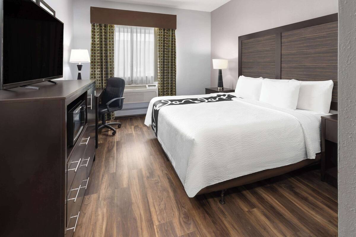Une chambre d’hôtel avec un grand lit, un bureau doté d’un téléviseur à écran plat, une chaise, une lampe de bureau et une fen être avec des rideaux.