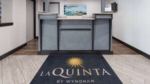 Un espace d’accueil doté d’un tapis noir orné du logo « LA QUINTA BY WYNDHAM », d’un comptoir de réception, d’un tableau représentant une cascade et d’un téléviseur fixé au mur.