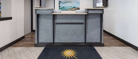 Zona de recepción con alfombra negra que muestra el logotipo “LA QUINTA BY WYNDHAM”, mostrador de recepción, pintura de una cascada y televisor montado.