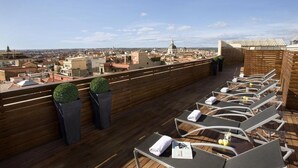 Une terrasse sur le toit, dotée de chaises longues et d’une plateforme en bois, offrant une vue panoramique sur la ville, avec ses bâtiments et sa structure en forme de dôme.