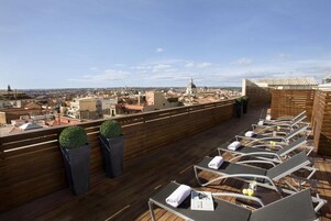 A rooftop terrace with lounge chairs and a wooden deck, overlooking a cityscape with buildings and a dome structure.