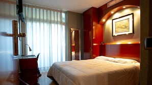 Habitación de hotel con cama, escritorio y silla. Hay una ventana con cortinas, un cuadro en la pared y una puerta.