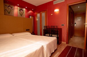 Ein Hotelzimmer mit zwei Betten, einem Holzkopfende, einer roten Akzentwand, einem Esstisch mit Stühlen und einer Tür zu einem weiteren Raum.