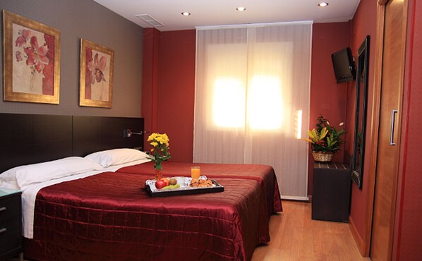 A hotel room with a large bed, a bedside table with a tray of fruit, a vase with flowers, and a television on the wall. - Hotel Villa De Barajas (Madrid)