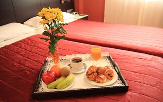 Ein Tablett mit Frühstücksangeboten, darunter Obst, Croissants und ein Glas Orangensaft, auf einem Bett mit roter Bettdecke.