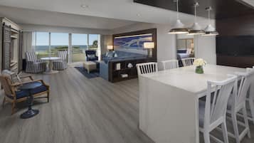 A modern hotel room with a large bed, a dining area, and a view of the ocean.