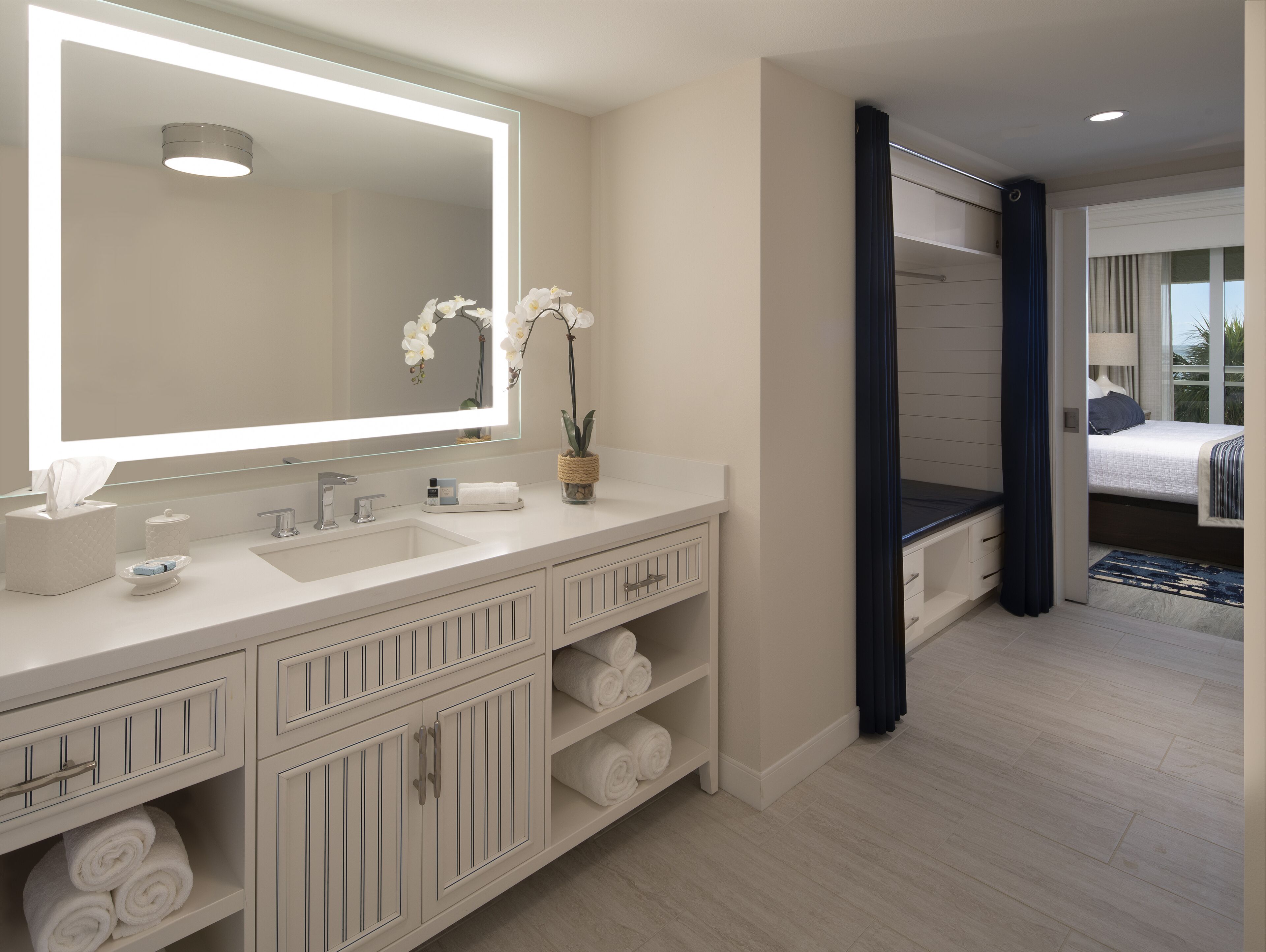 A modern bathroom with a large mirror, white fixtures, and a vase with white flowers.