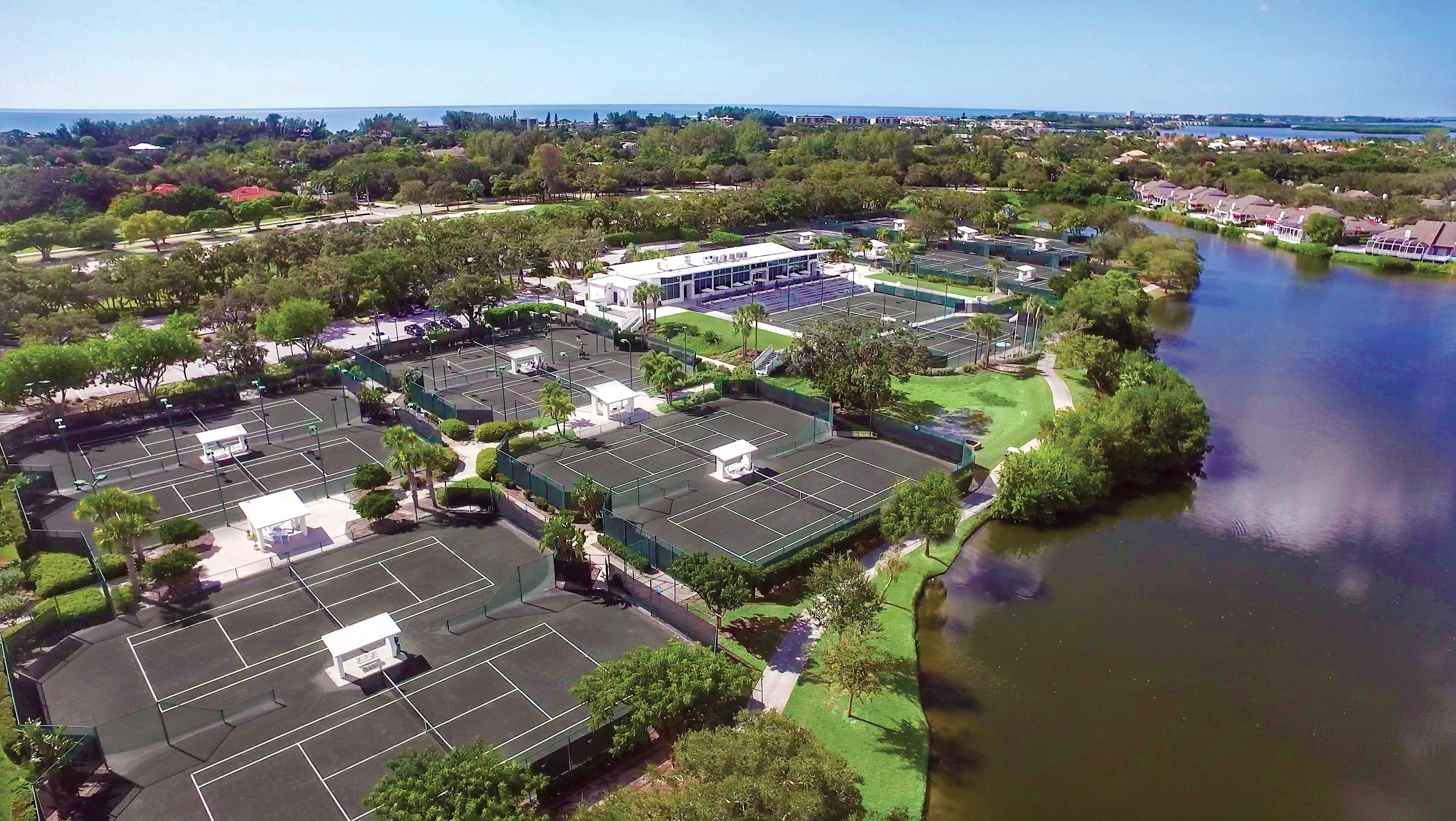 An aerial view of a residential area with multiple tennis courts, a river, and surrounding greenery.