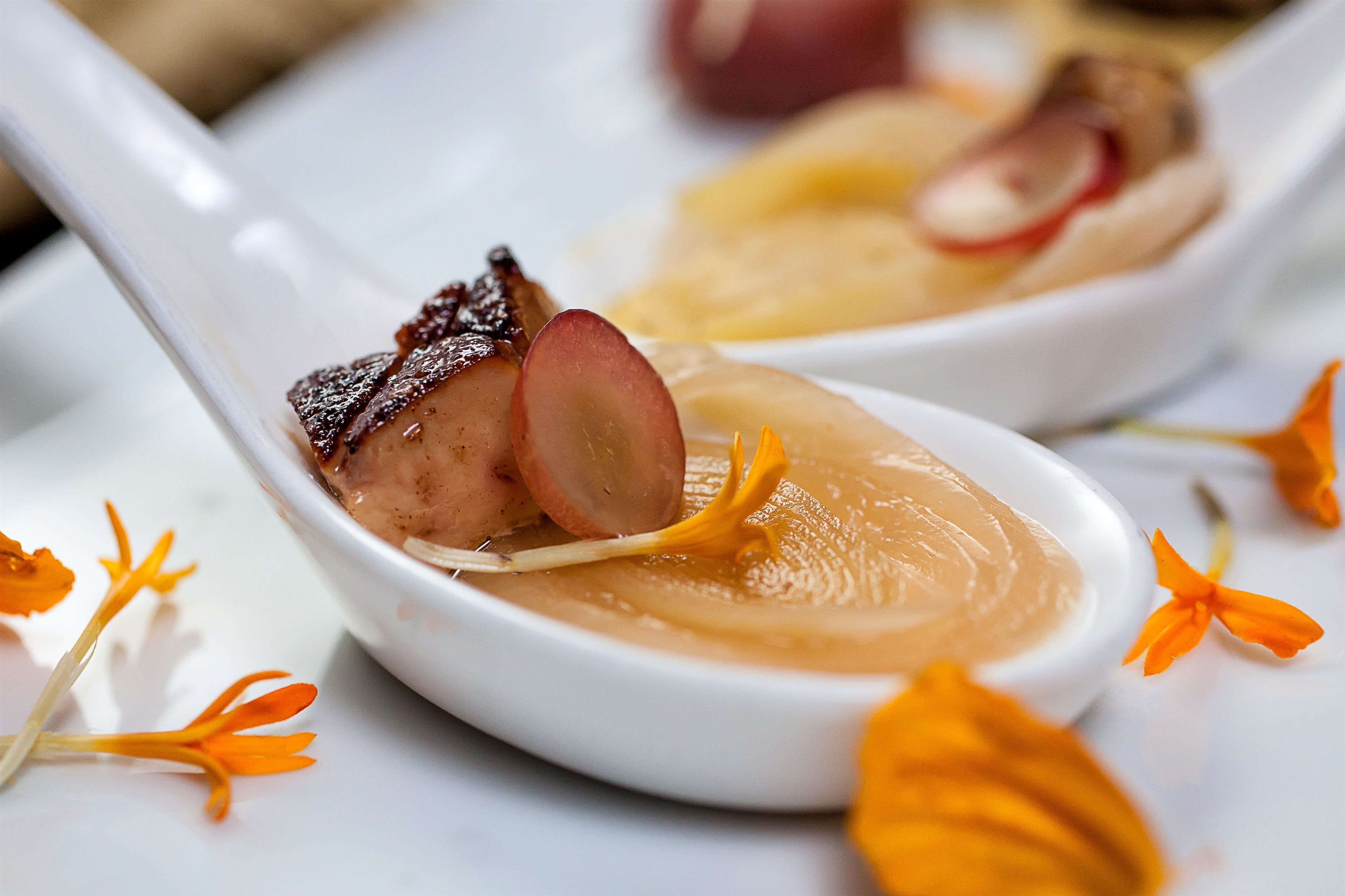 A close-up of a white ceramic spoon with a creamy soup and a piece of meat, surrounded by orange flowers.
