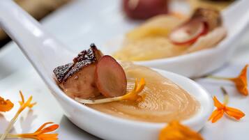 A close-up of a white ceramic spoon with a creamy soup and a piece of meat, surrounded by orange flowers.