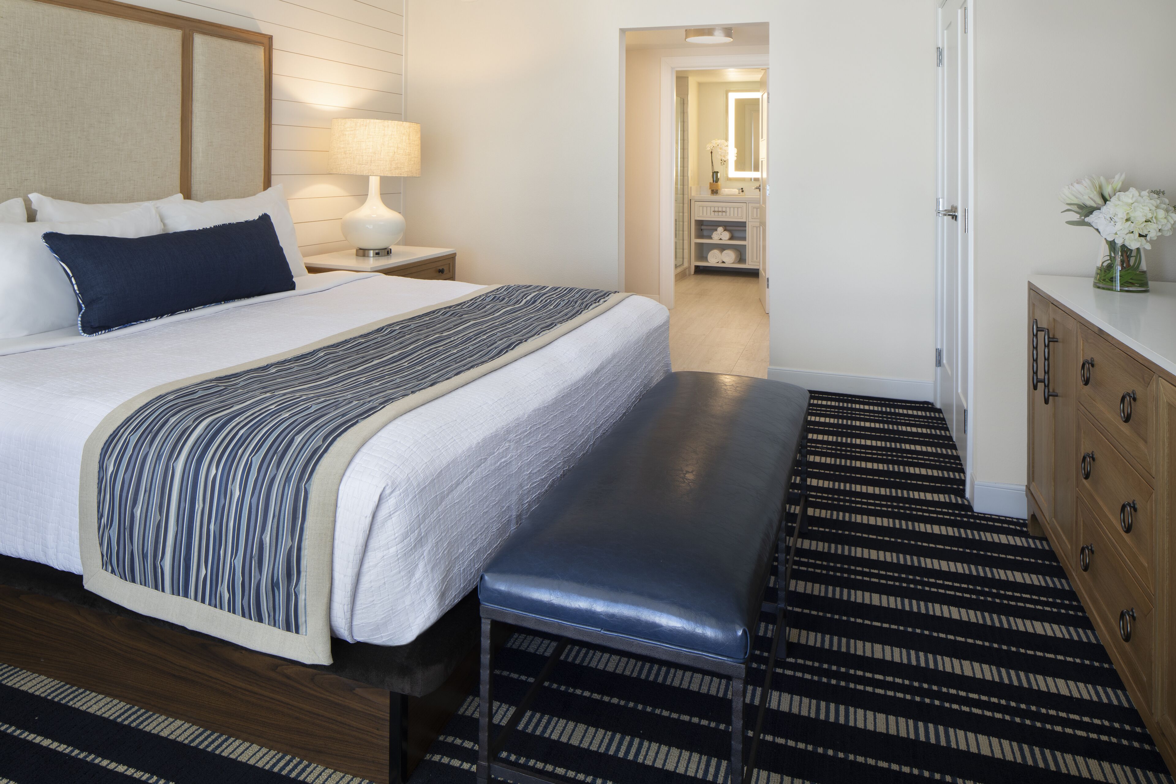 Habitación de hotel con una cama grande, un banco, un aparador de madera y un baño visible a través de una puerta abierta.