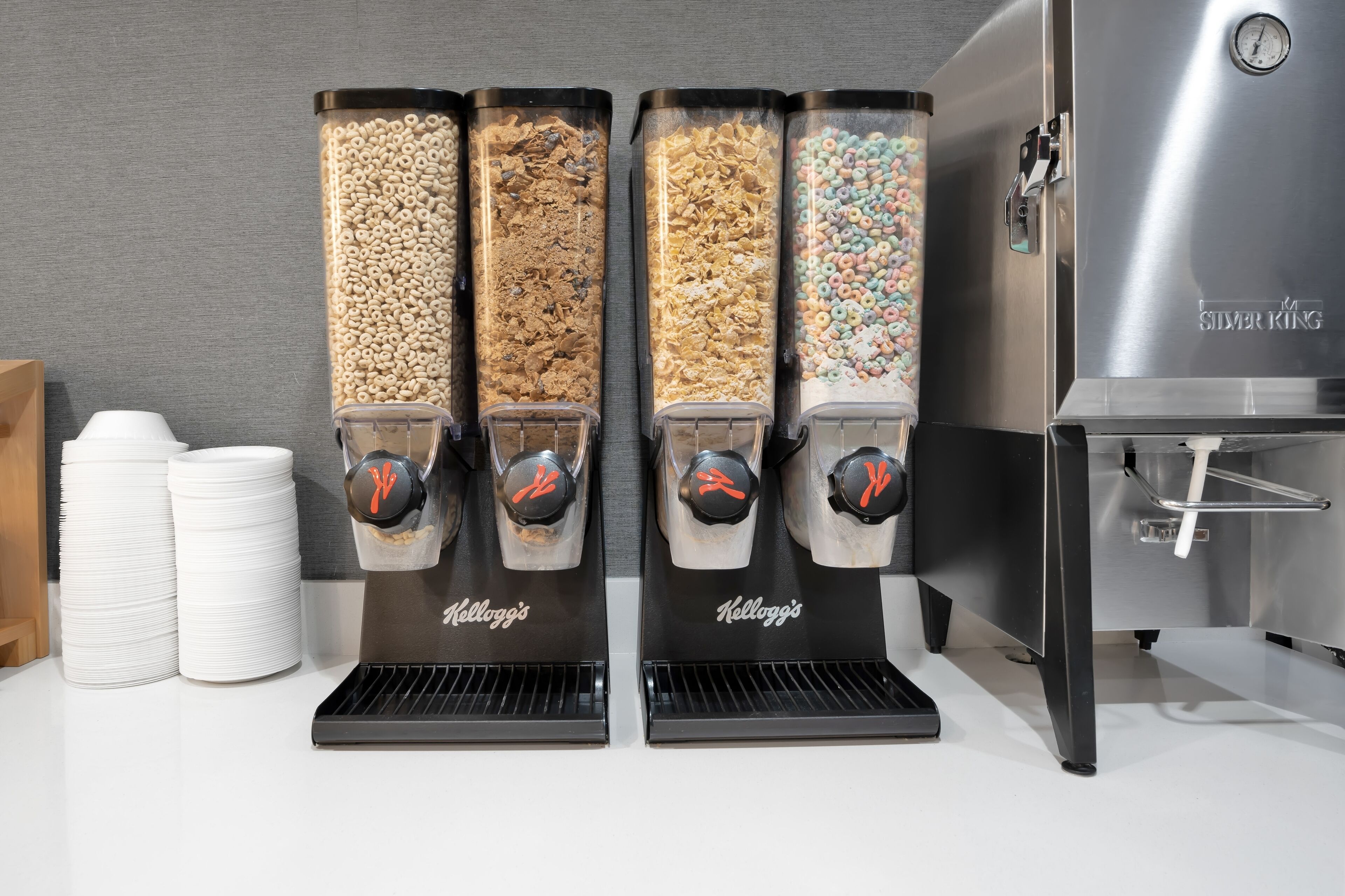 Cuatro dispensadores de cereal con distintos sabores, una pila de vasos blancos y una máquina de acero inoxidable con la etiqueta "Silver King".