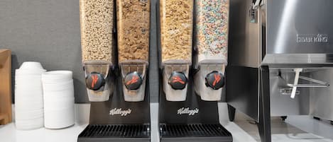 Cuatro dispensadores de cereal con distintos sabores, una pila de vasos blancos y una máquina de acero inoxidable con la etiqueta "Silver King".