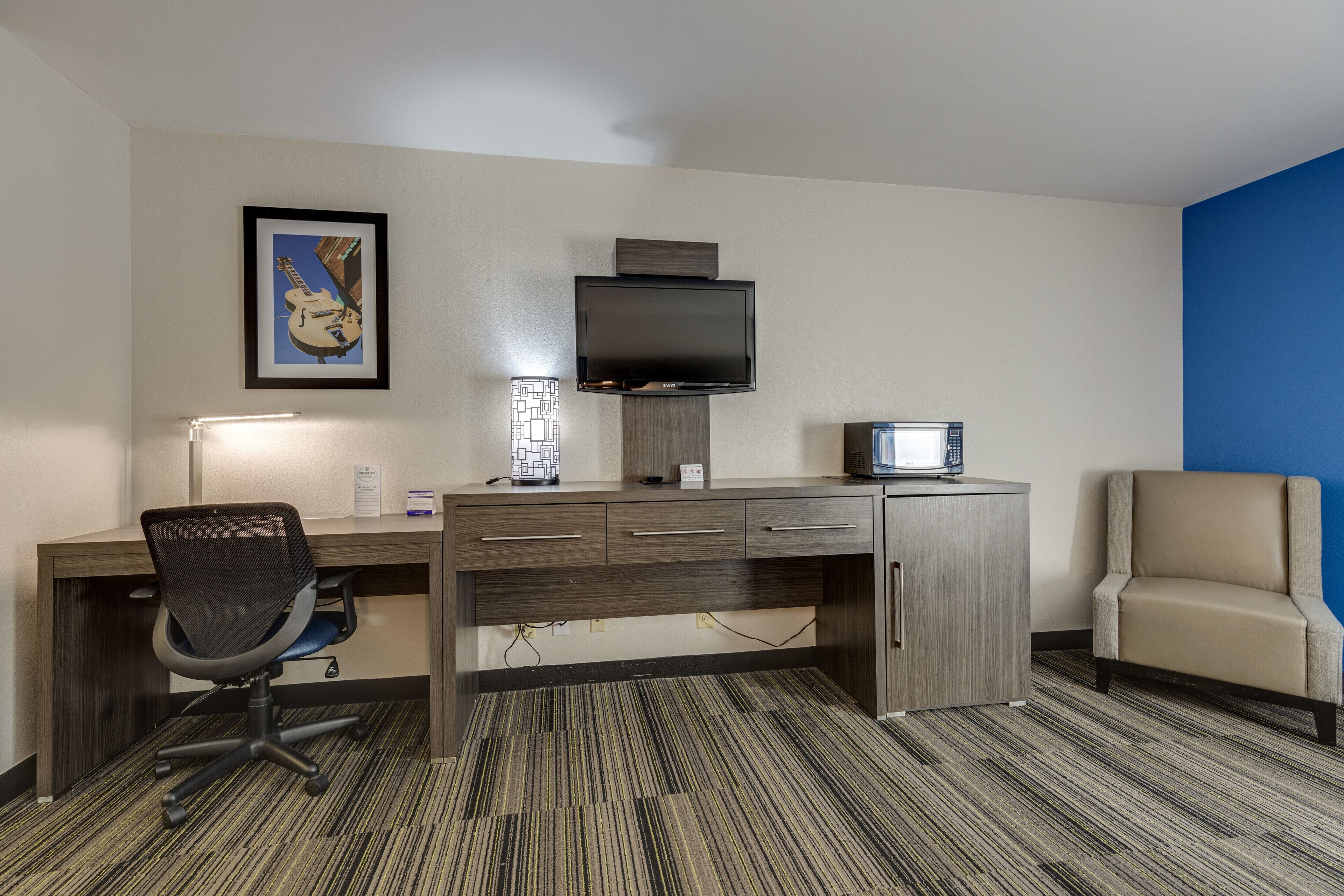 A hotel room with a desk, chair, television, and a blue accent wall.