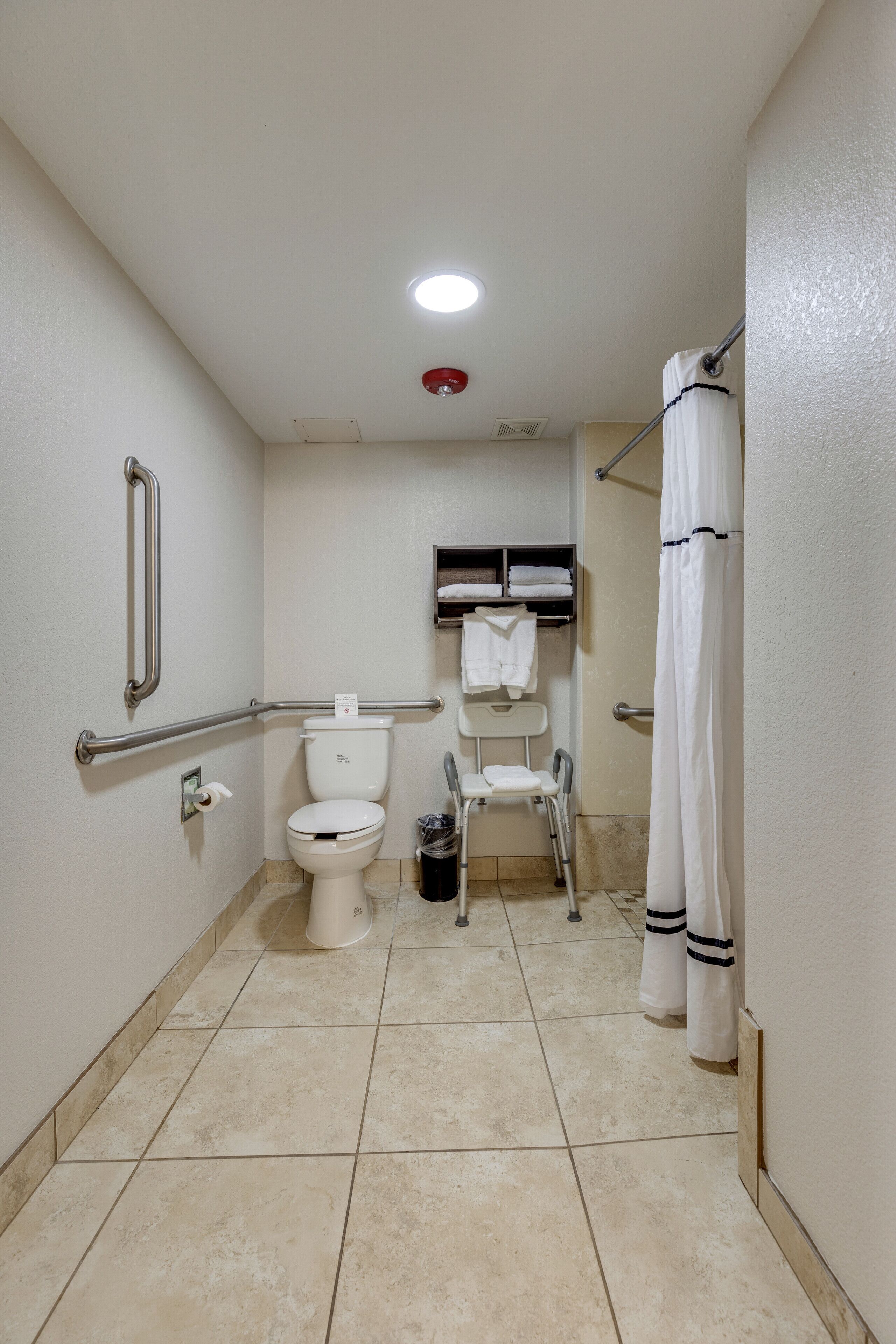 Un baño pequeño y limpio con inodoro, barras de apoyo, cortina de ducha y una silla pequeña.