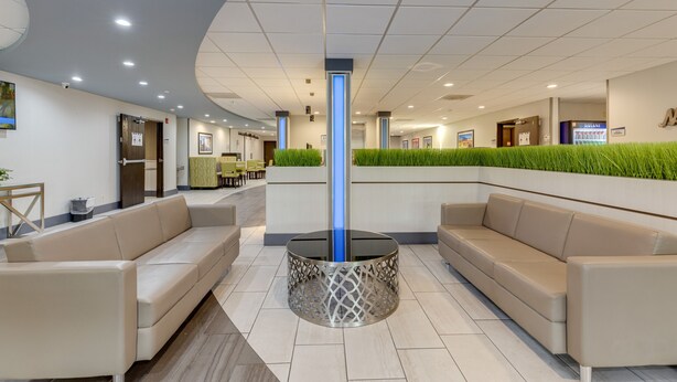 Un lobby moderno con una columna central de vidrio, sofás color beige y una pared verde con plantas.