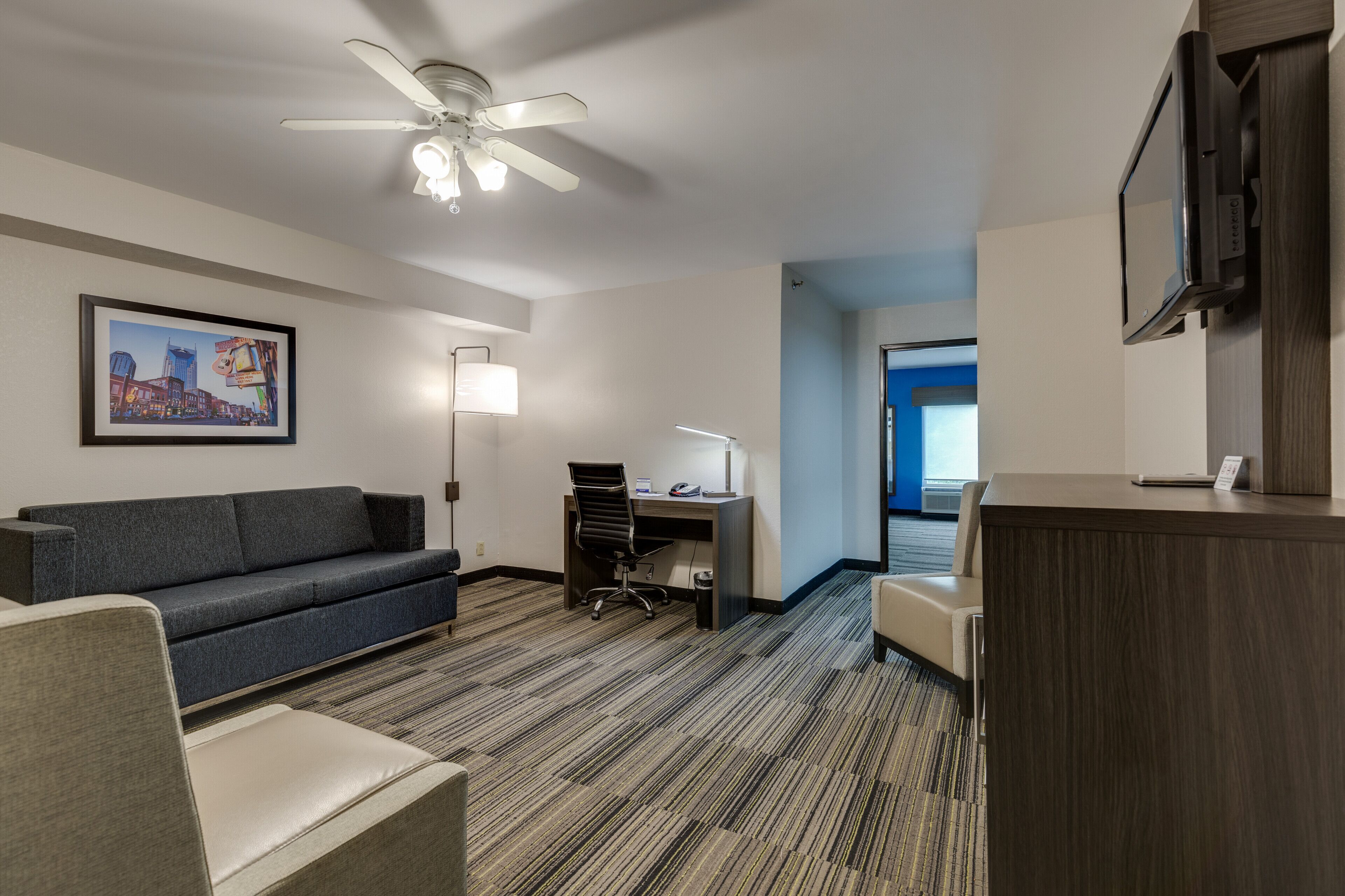 Habitación de hotel con un sofá gris, un escritorio con silla, ventilador de techo, televisión e imagen de un paisaje urbano enmarcada en la pared.