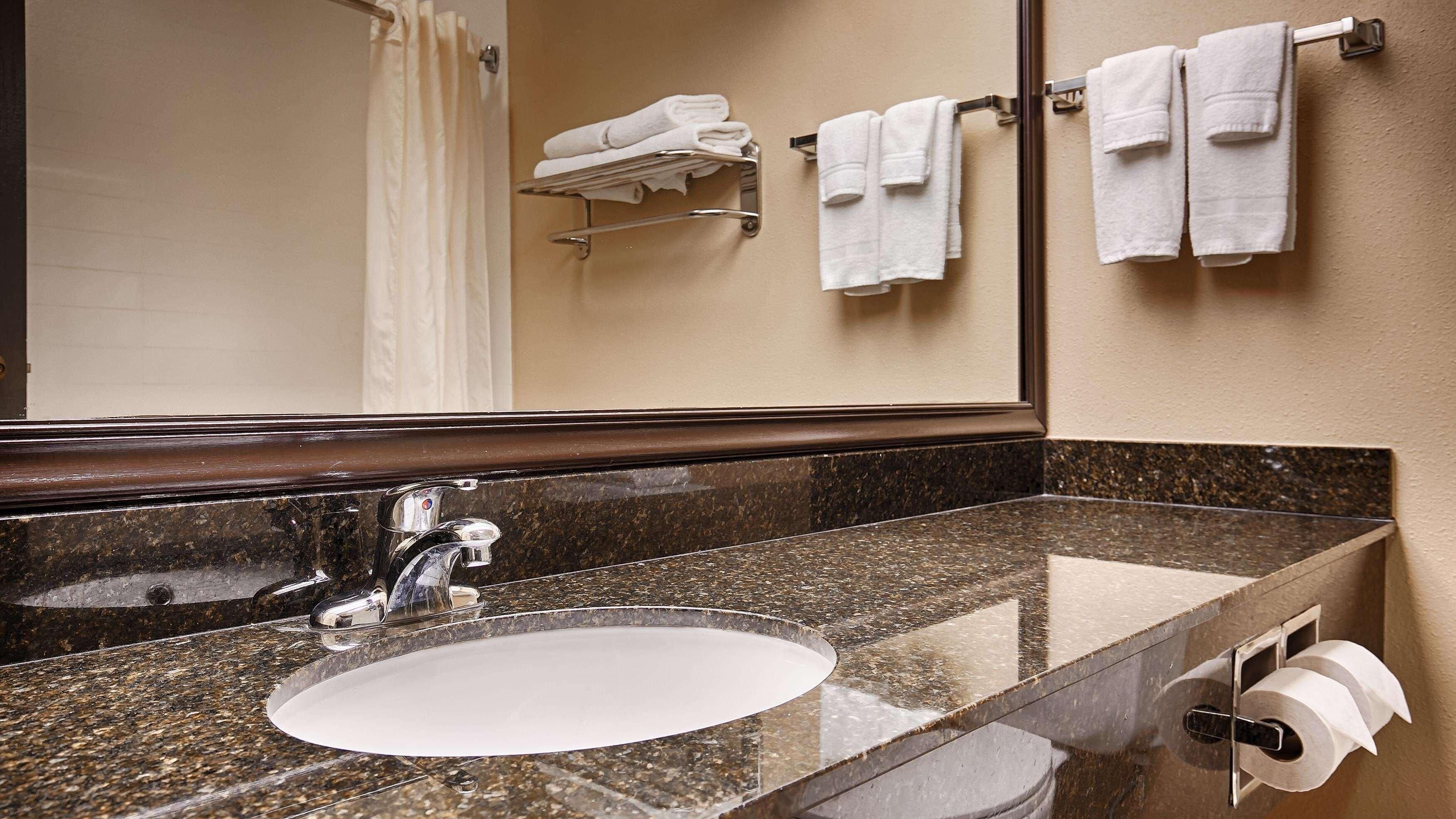 A bathroom with a granite countertop sink, a chrome faucet, a towel rack with folded towels, and a toilet paper holder.