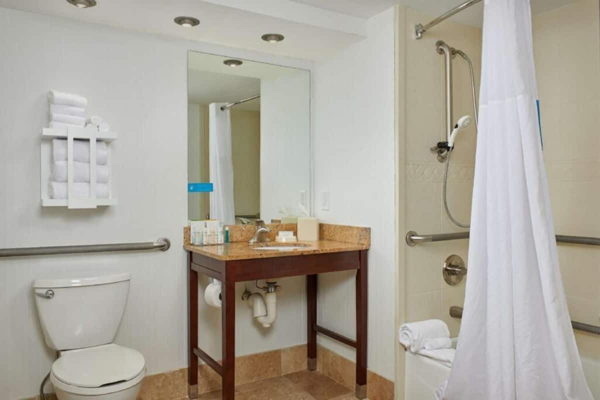 A bathroom with a toilet, sink, mirror, towel rack, shower, and grab bars.