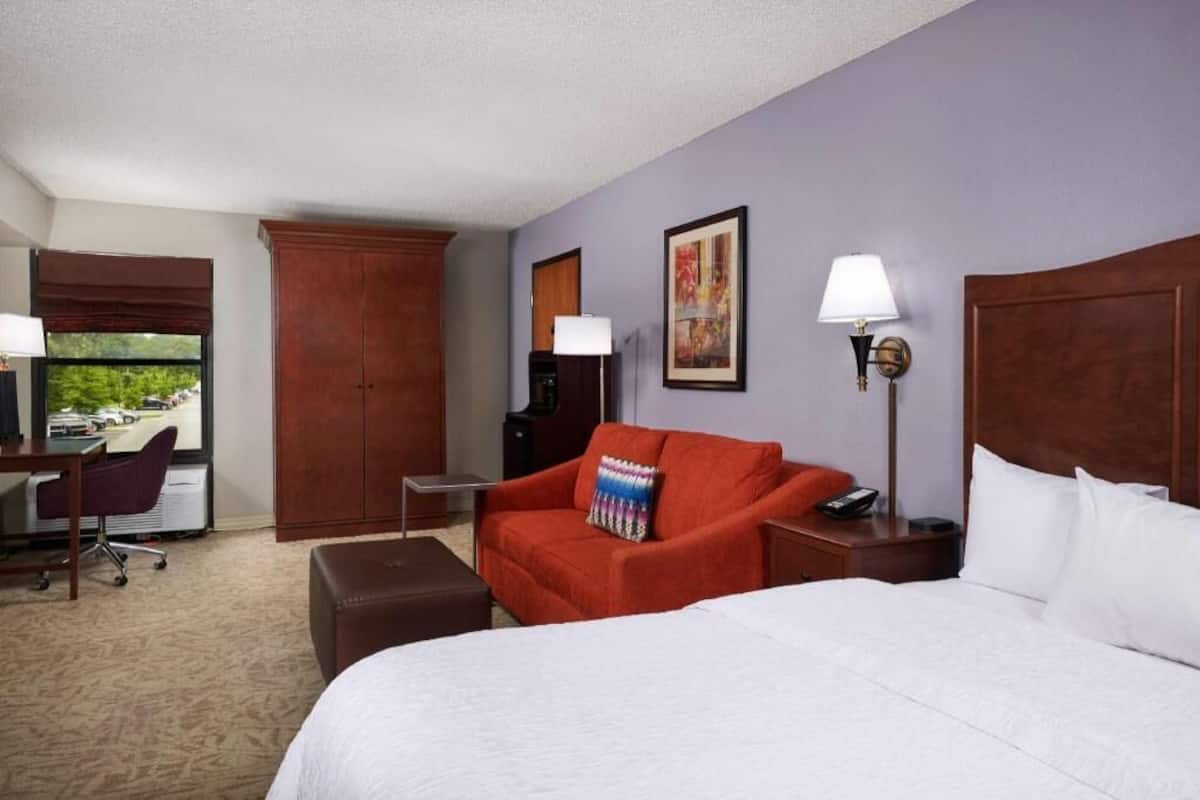 A hotel room with a bed, a red armchair, a desk, a lamp, and a painting on the wall.