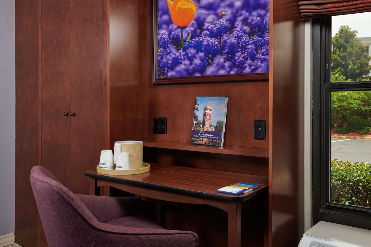 A hotel room with a wooden wardrobe, a purple chair, a small table with a tray of toiletries, a framed picture of a flower, and a framed book.