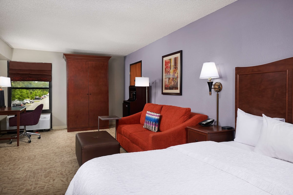 A hotel room with a large bed, a red armchair, a desk, and a view of the outdoors.