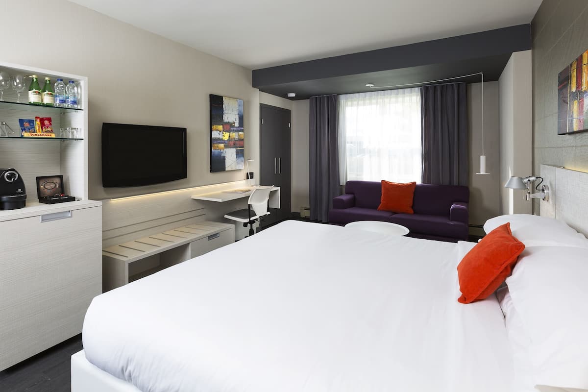 Una habitación de hotel moderna con una cama grande, un escritorio con una silla, un televisor de pantalla plana, un sofá y un minibar.