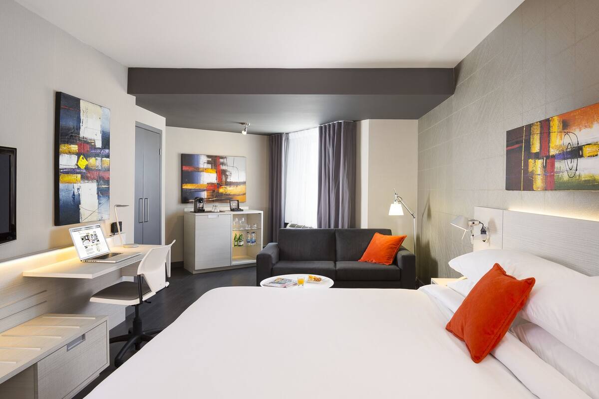 Una habitación de hotel moderna con una cama grande, un escritorio con una computadora portátil, un sofá y un televisor.
