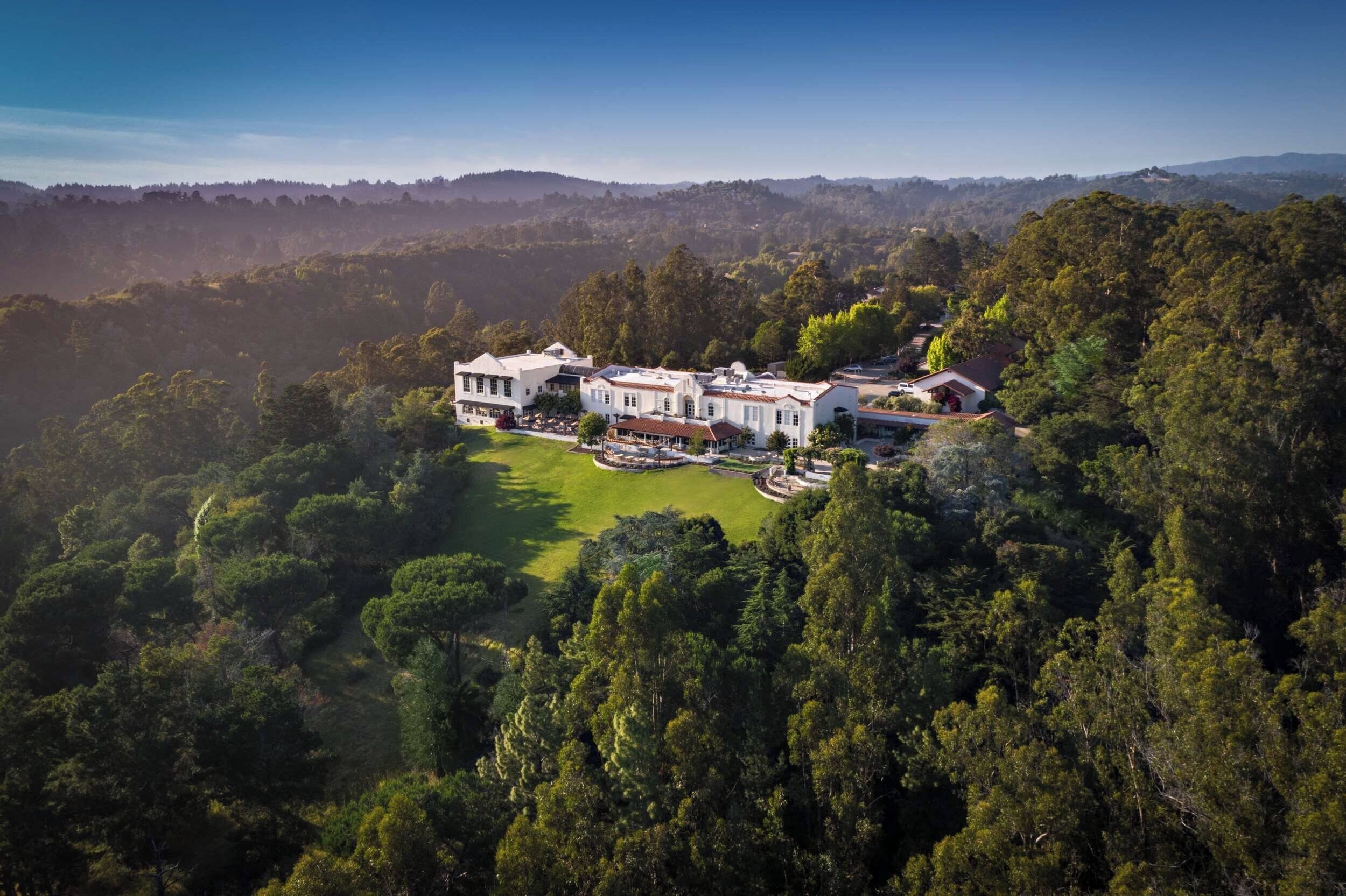A large, multi-story estate with a well-maintained lawn and a variety of buildings, surrounded by dense forest.