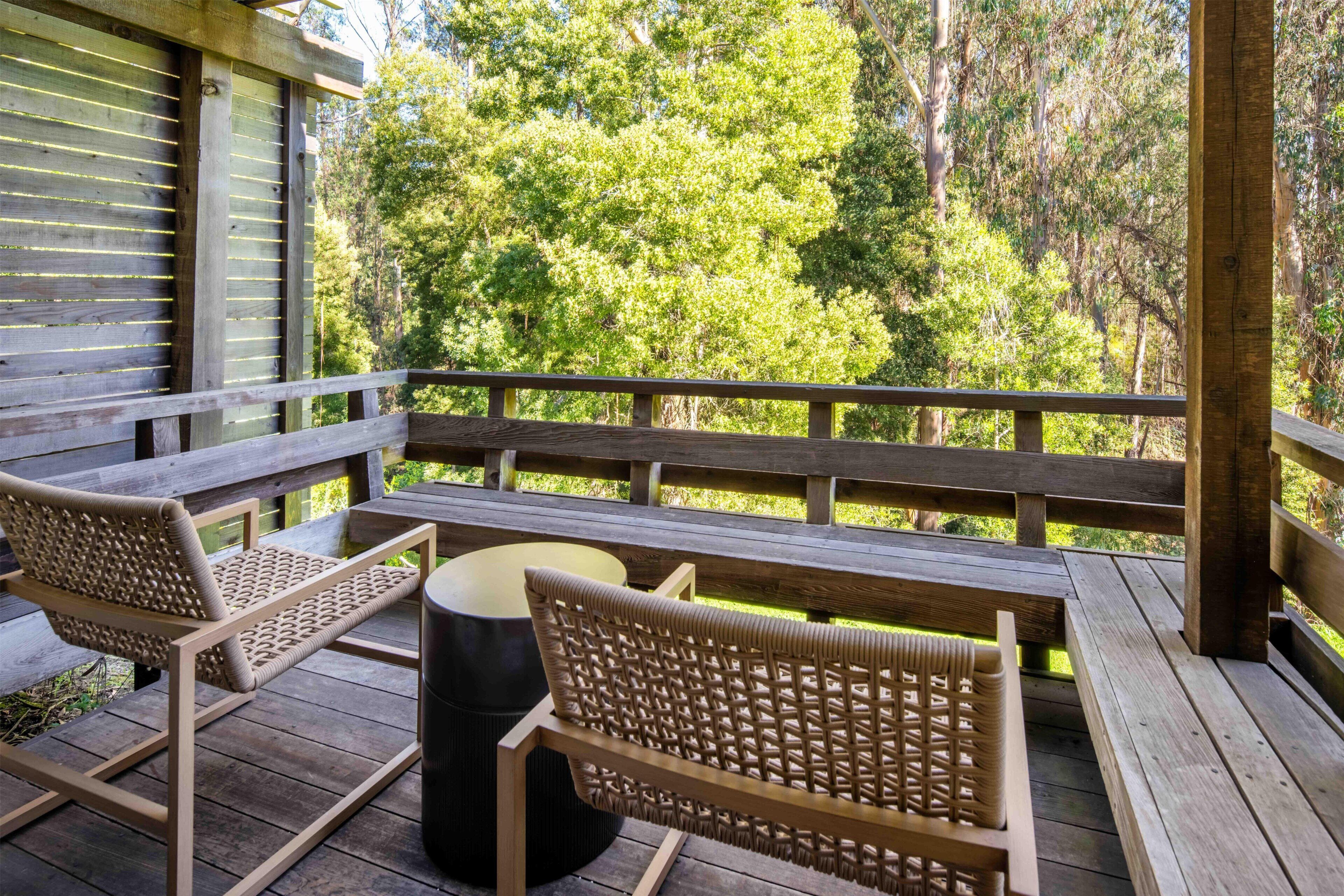 A wooden deck with a table and two wicker chairs, overlooking a forested area.