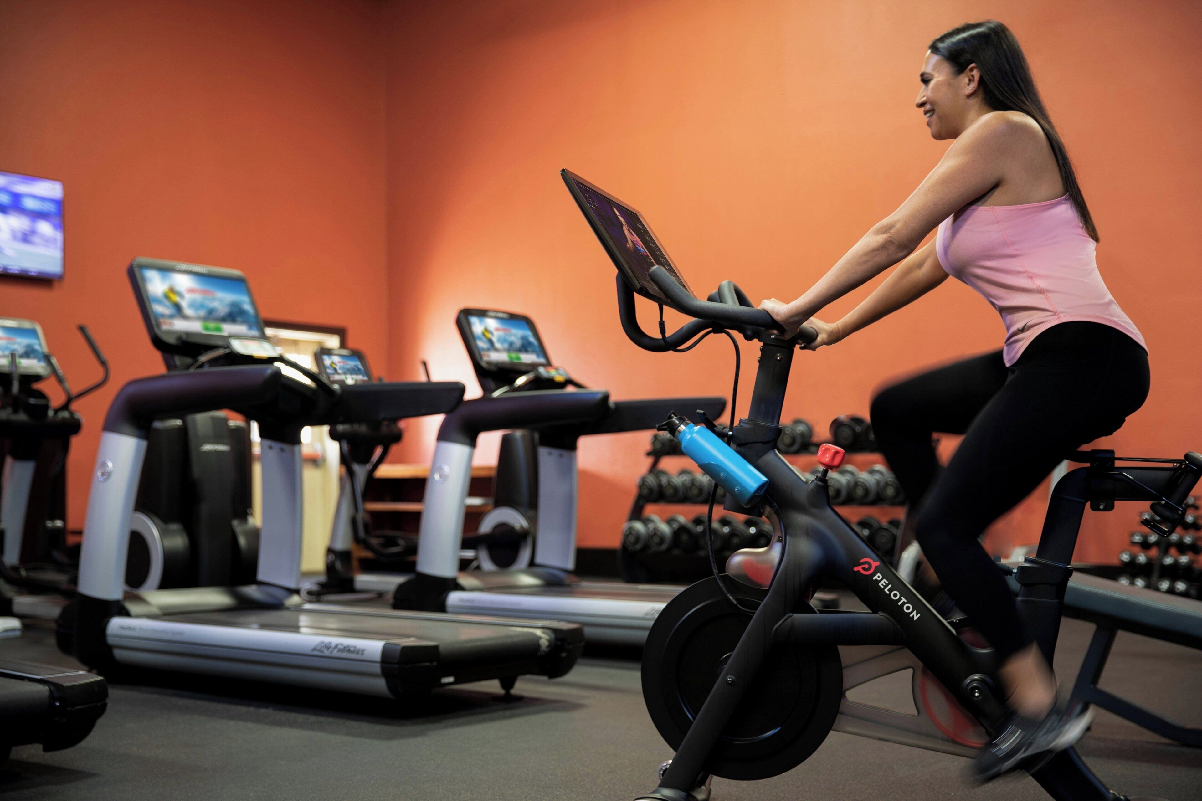 Una persona usando una bicicleta estática en un gimnasio con cintas de correr y mancuernas al fondo.