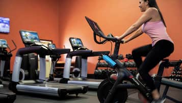 Una persona usando una bicicleta estática en un gimnasio con cintas de correr y mancuernas al fondo.
