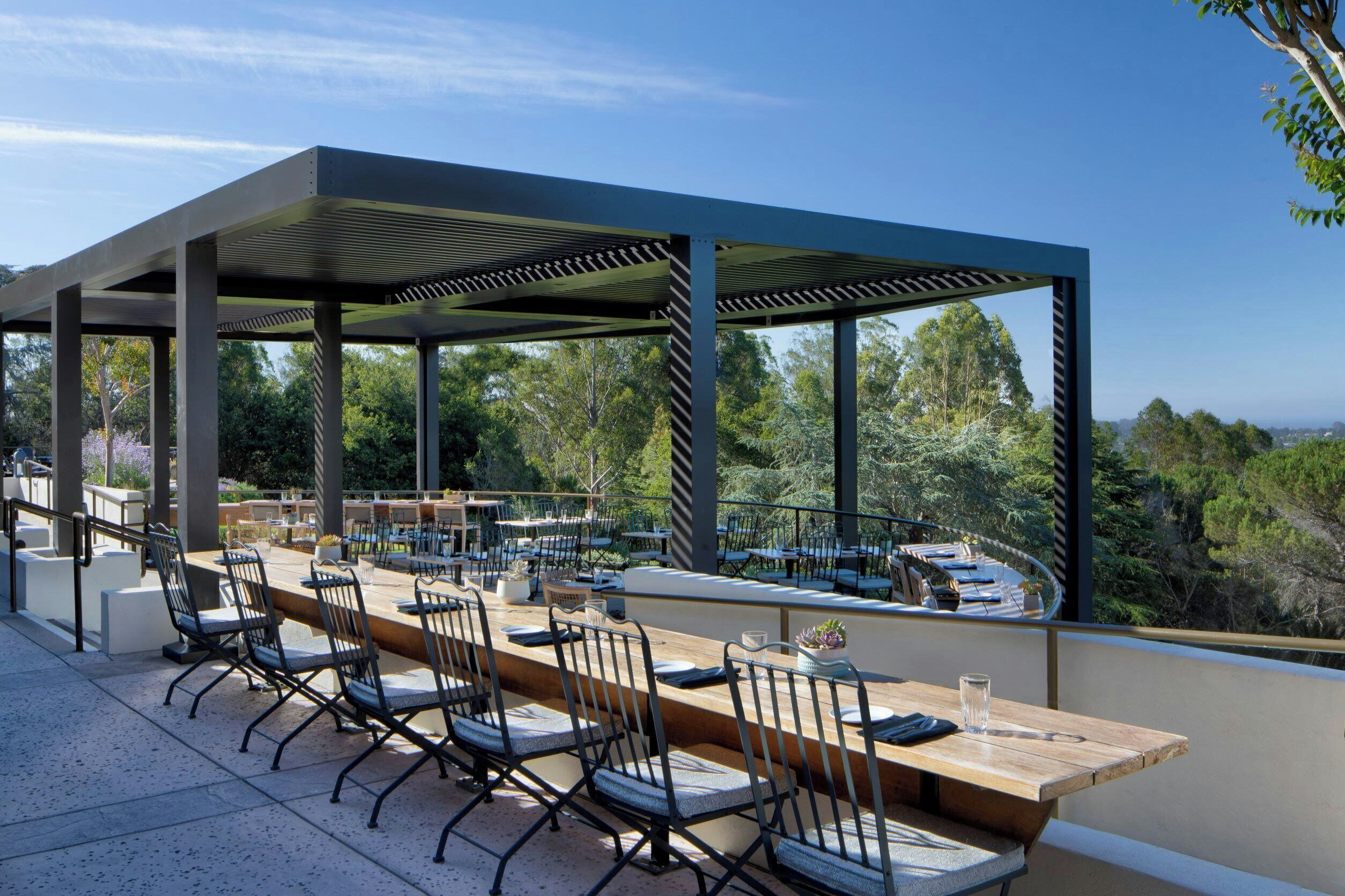 An outdoor dining area with a long wooden table and chairs, surrounded by a covered pergola with a view of the forest.