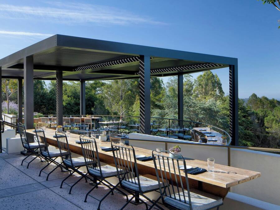 An outdoor dining area with a long wooden table and chairs, surrounded by a covered pergola with a view of the forest.