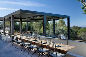 Un espace de restauration en plein air avec une longue table et des chaises en bois, entouré d’une pergola couverte offrant une vue sur la forêt.