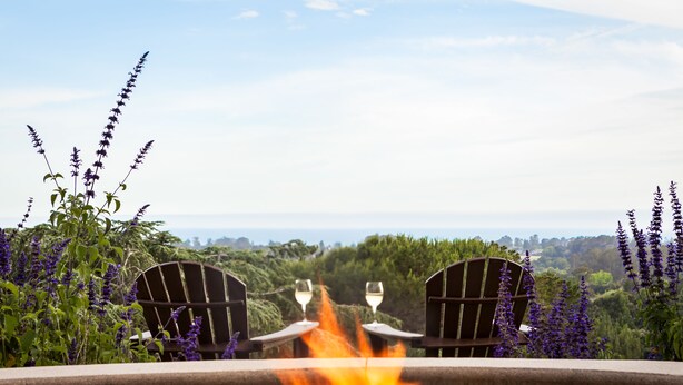 A patio with two chairs and a table, a fire in the center, surrounded by greenery and a view of the landscape. - Chaminade Resort & Spa (Santa Cruz)