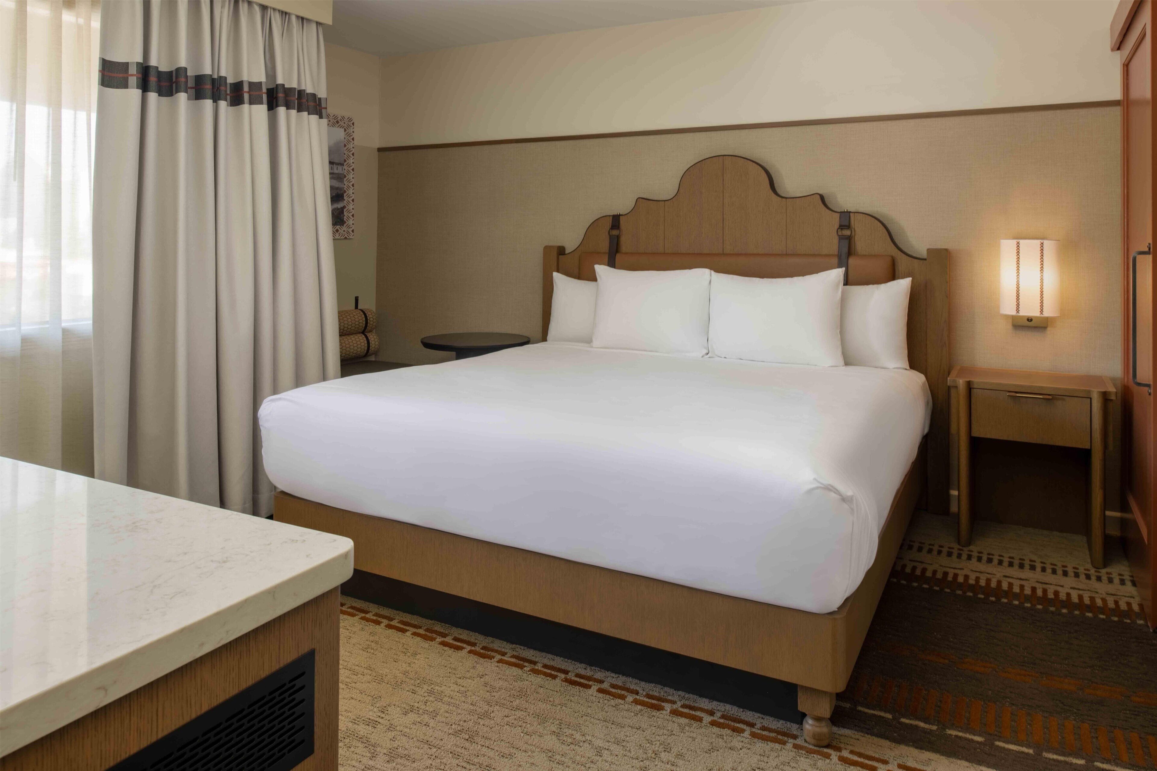 A hotel room with a bed, bedside table, lamp, and a view of the outside through curtains.
