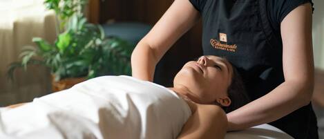 Une personne allongée sur un lit recouvert d’une couverture blanche reçoit un massage du dos d’une autre personne qui porte un t-shirt noir orné d’un logo.
