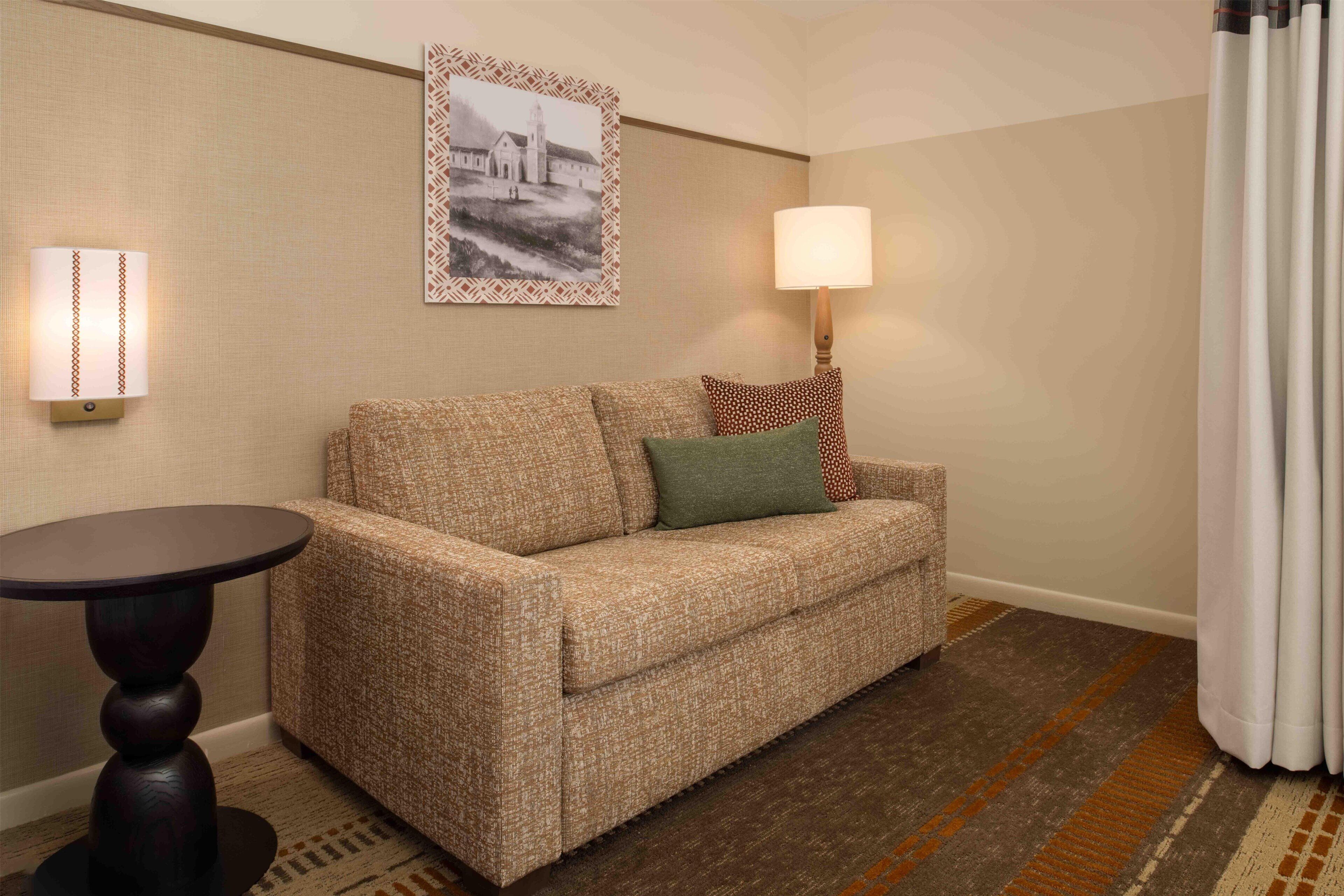 A hotel room with a patterned sofa, a small round table, a wall-mounted lamp, and a framed picture on the wall.