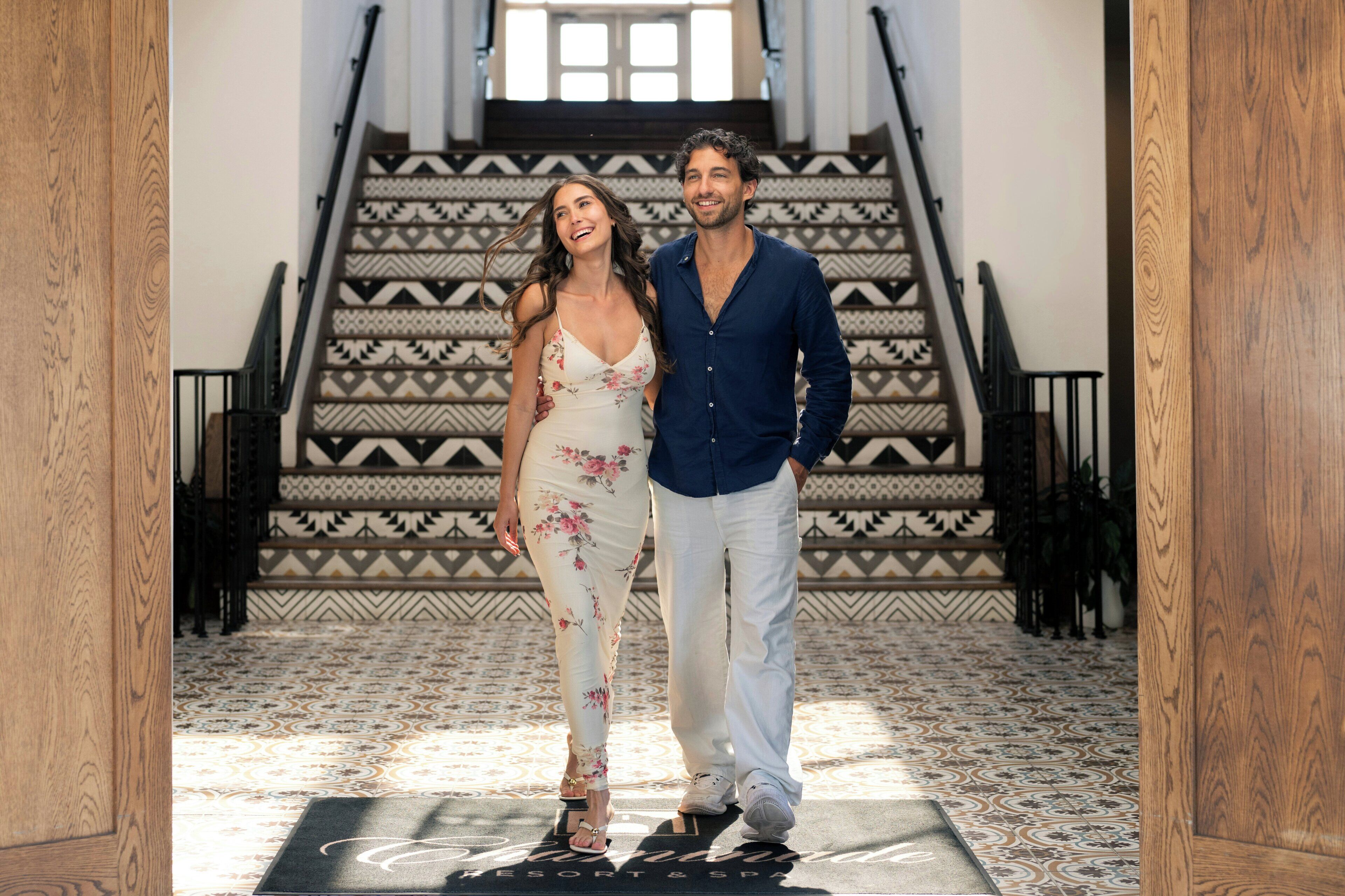 A man and a woman standing in a hallway with a patterned floor and a staircase with geometric designs.