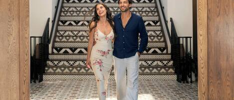 A man and a woman standing in a hallway with a patterned floor and a staircase with geometric designs.