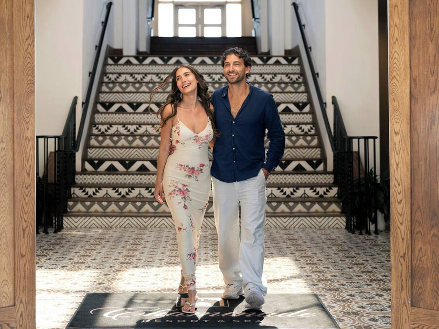 A man and a woman standing in a hallway with a patterned floor and a staircase with geometric designs.