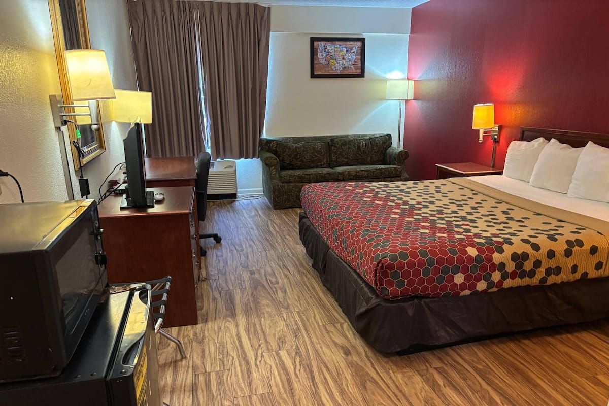 A hotel room with a bed, a desk with a computer, a TV, a couch, and a window with curtains.