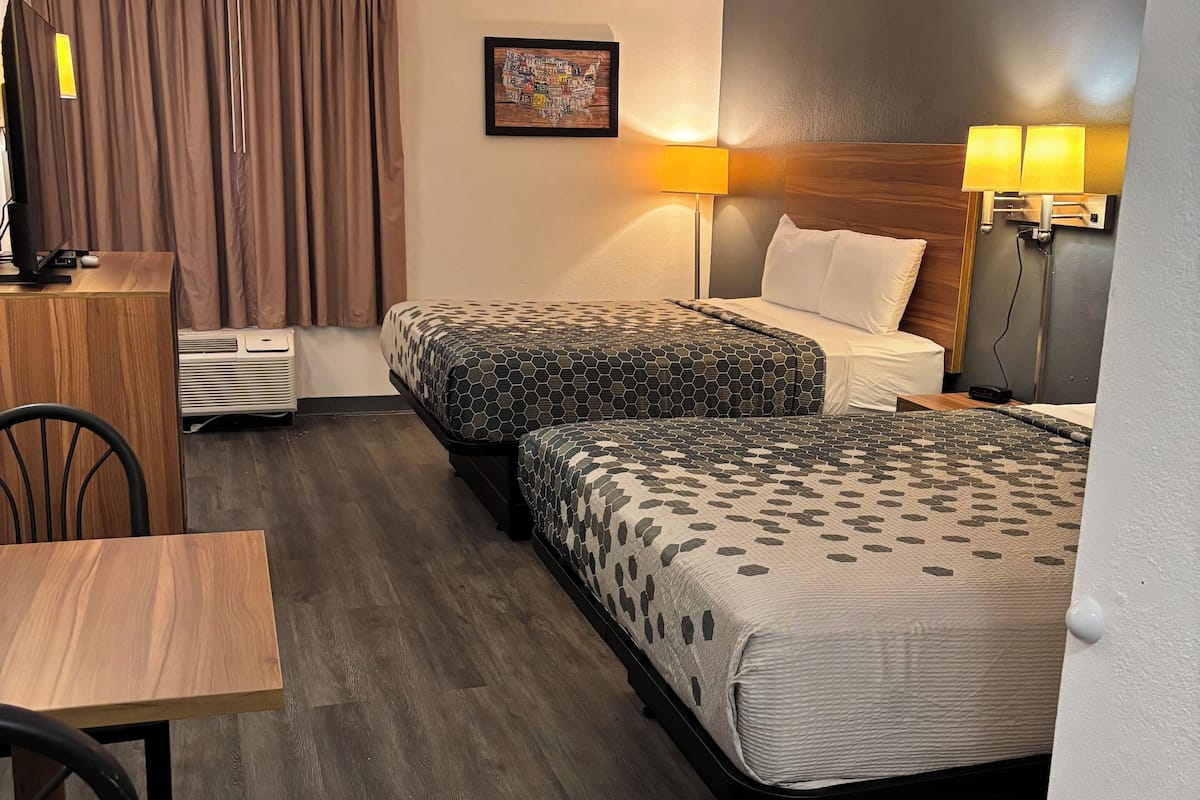 A hotel room with two beds, a desk, a chair, a desk lamp, and a wall-mounted TV.