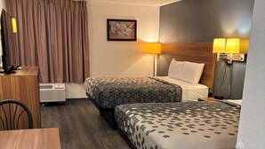 Habitación de hotel con dos camas, un escritorio, una silla, una lámpara de escritorio y un televisor montado en la pared.