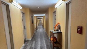 Un pasillo de hotel con suelo de madera, paredes amarillas y puertas a ambos lados. A la derecha, hay una pequeña mesa con un adorno.