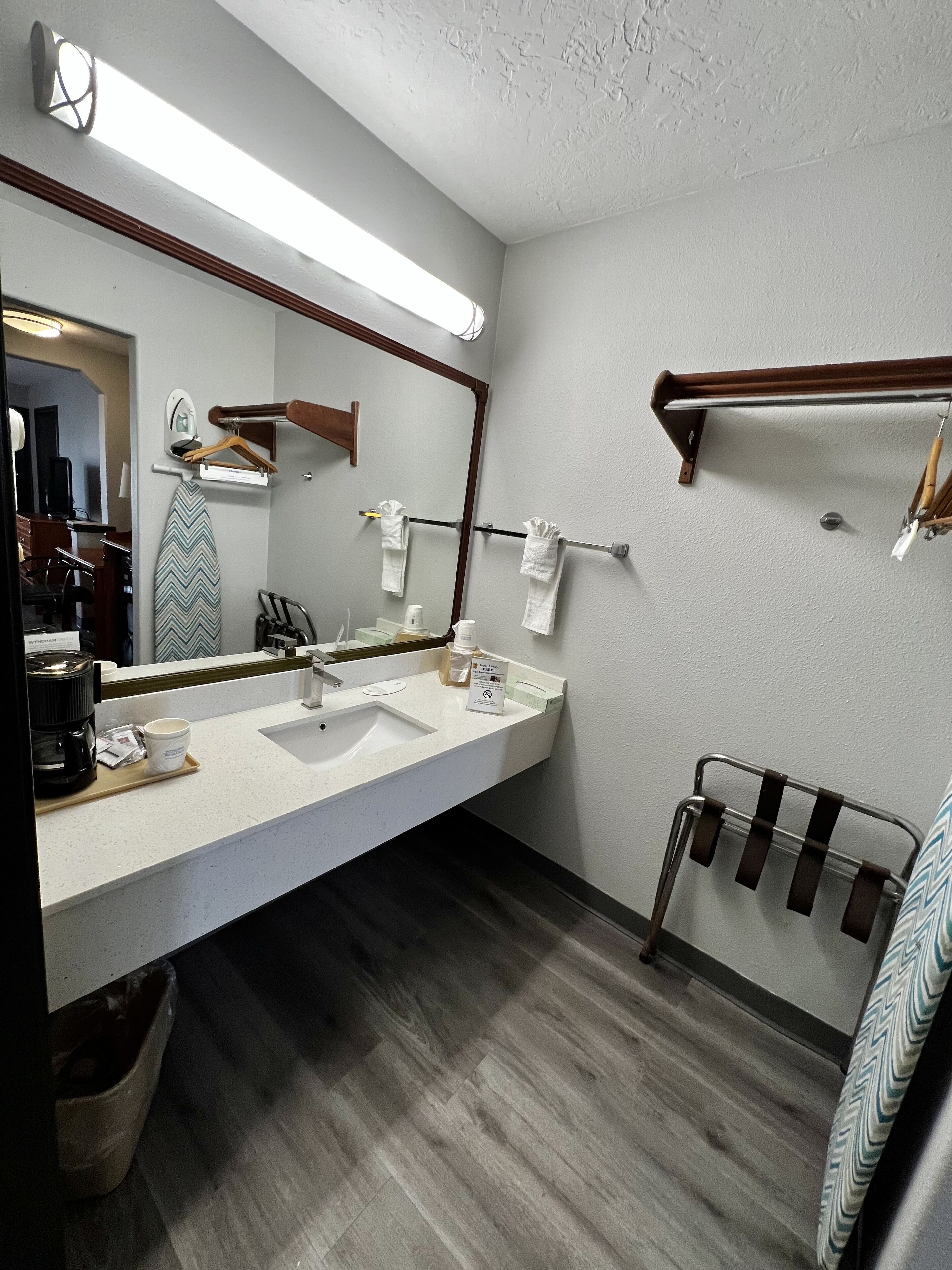 A hotel bathroom with a large mirror, two sinks, a towel rack, and a wooden floor.