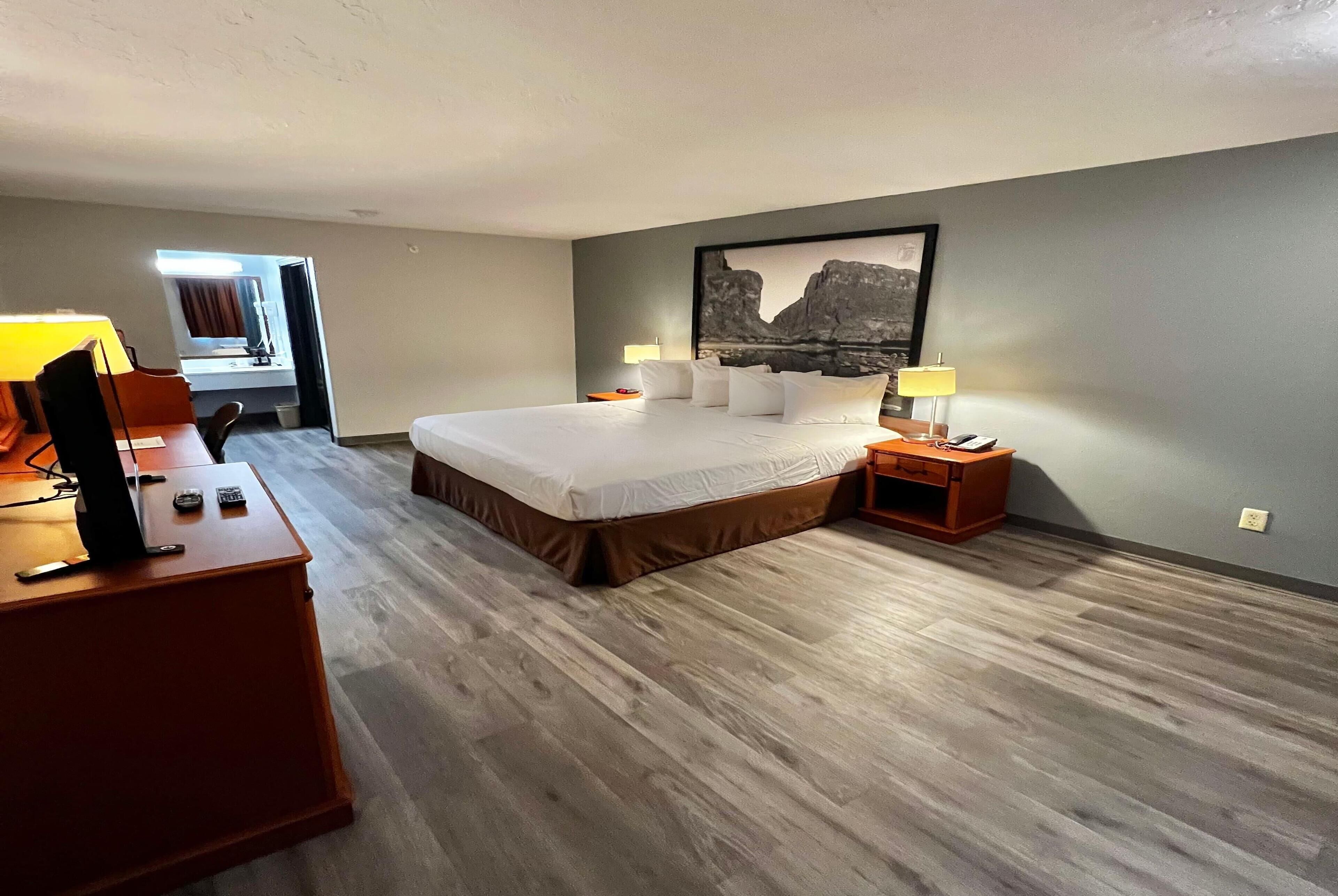 A hotel room with a large bed, a desk with a lamp, a nightstand, and a framed picture on the wall.