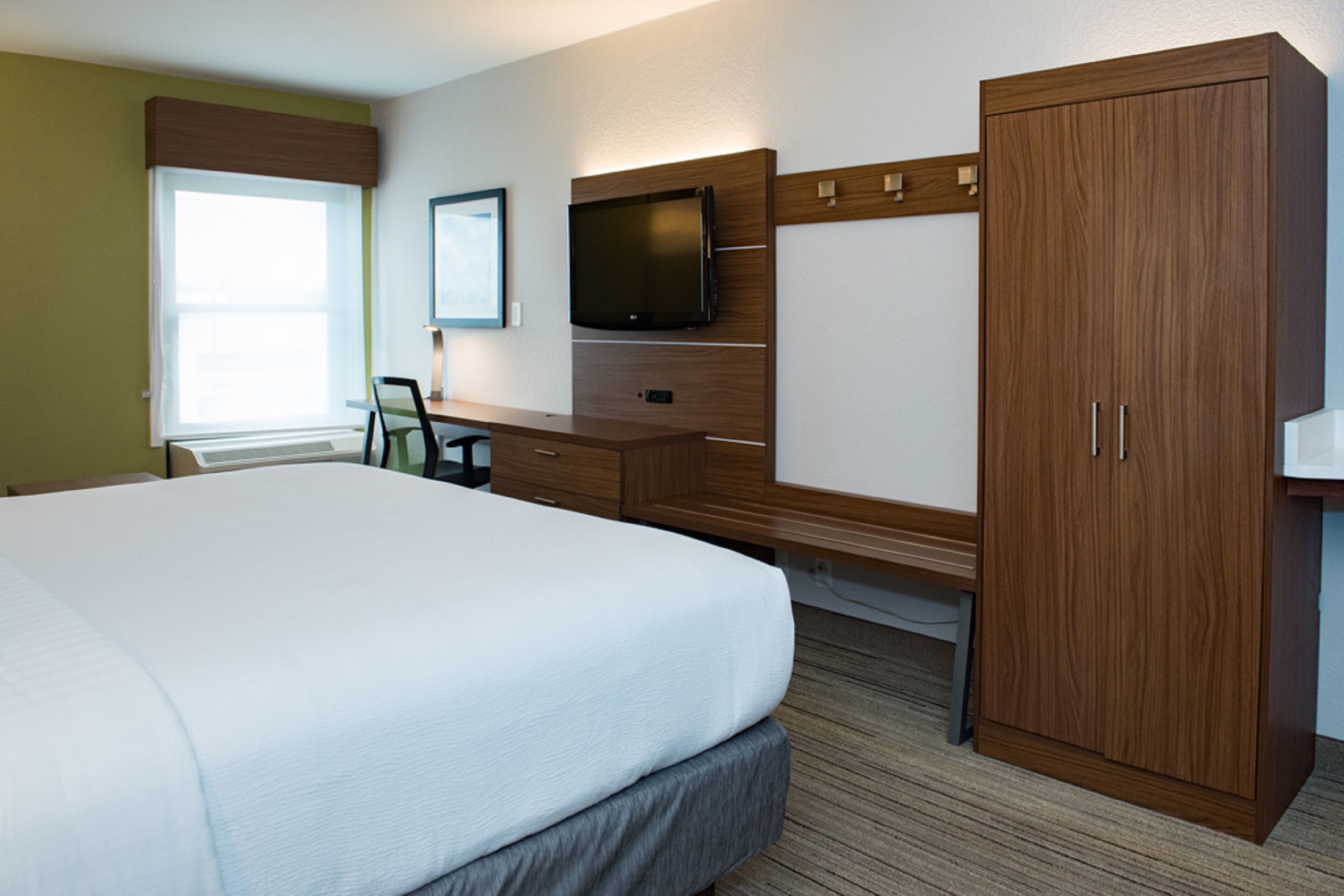 Une chambre d’hôtel dotée d’un grand lit, d’une armoire en bois, d’un bureau avec une chaise, d’un téléviseur à écran plat fixé au mur et d’une fenêtre laissant entrer la lumière naturelle.