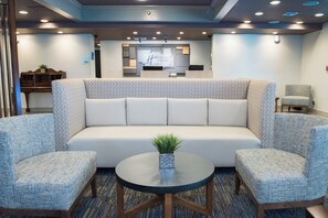A modern hotel lobby with a sofa, two armchairs, a small round table with a plant, and a television screen in the background.